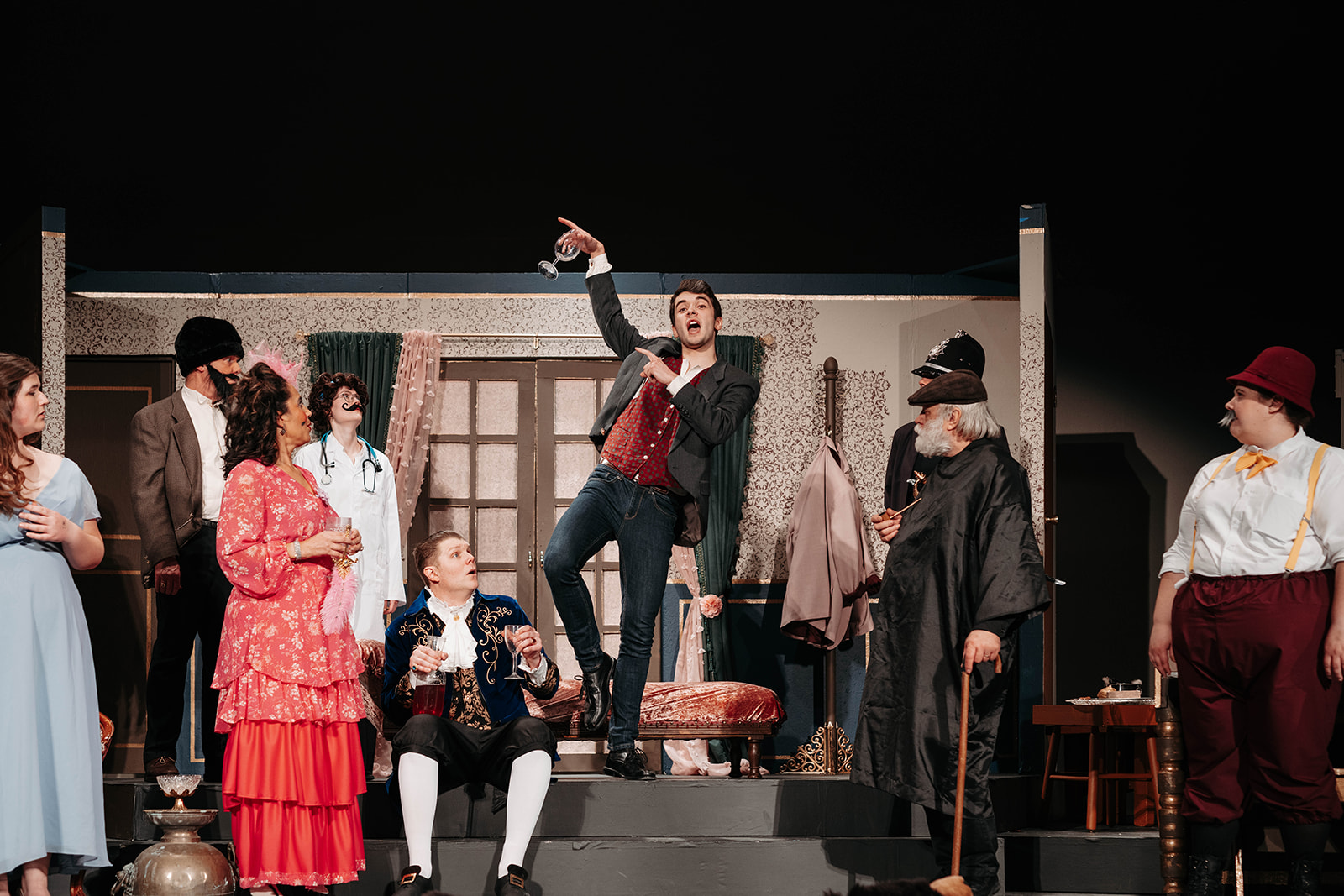 Rosetown Playhouse A dramatic scene showing a performer standing triumphantly on furniture with one arm raised, surrounded by other cast members in period dress including elaborate gowns and formal attire.