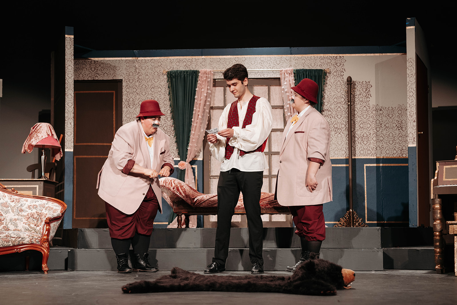 Rosetown Playhouse Three actors on stage, with two men in beige suits and burgundy hats presenting something to a young man in a red vest and white shirt, with a fur rug visible on the floor.
