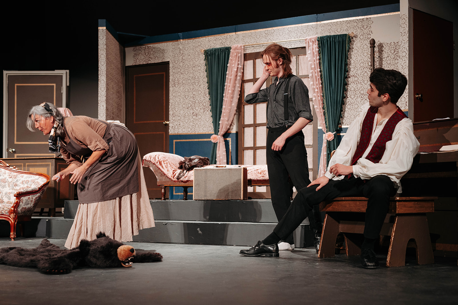 Rosetown Playhouse Scene showing three performers including an elderly woman in period dress bending over, a person in dark clothing, and a young man in a vest seated on a wooden chair.