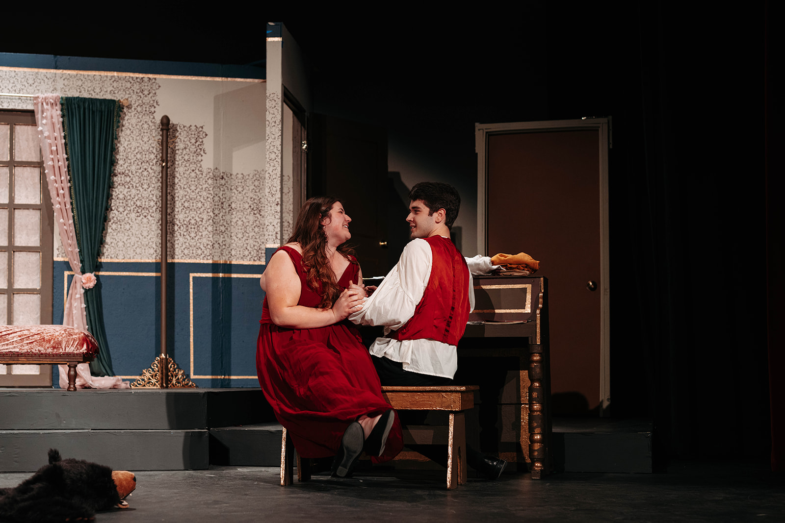Rosetown Playhouse A romantic scene with two performers in matching red vests sitting close together on a bench, gazing at each other against the floral-papered set.