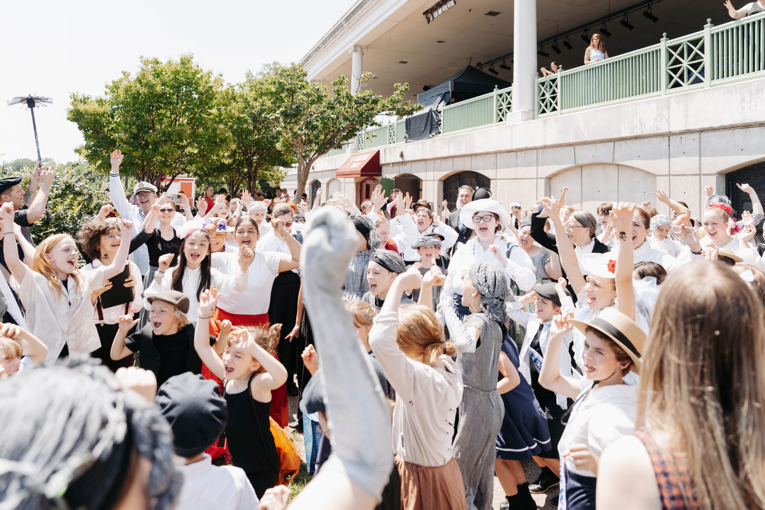 Large group of children in vintage early 1900s costumes with raised arms celebrating outdoors in front of a white colonial-style building with green balcony railings. Children wear period attire including sailor outfits, white blouses, long skirts, suspenders, and caps, appearing to be part of a theatrical performance or historical reenactment.