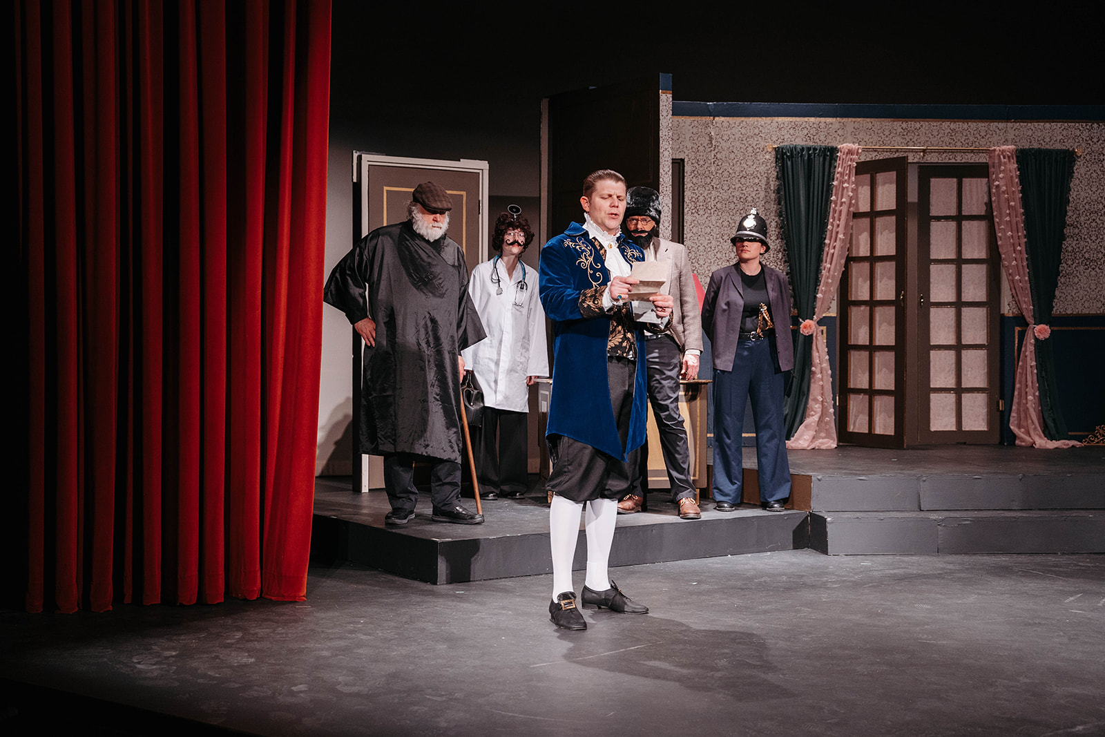 Rosetown Playhouse A formal scene with an actor in an elaborate blue period coat reading from a paper center stage, flanked by other characters in various period costumes.