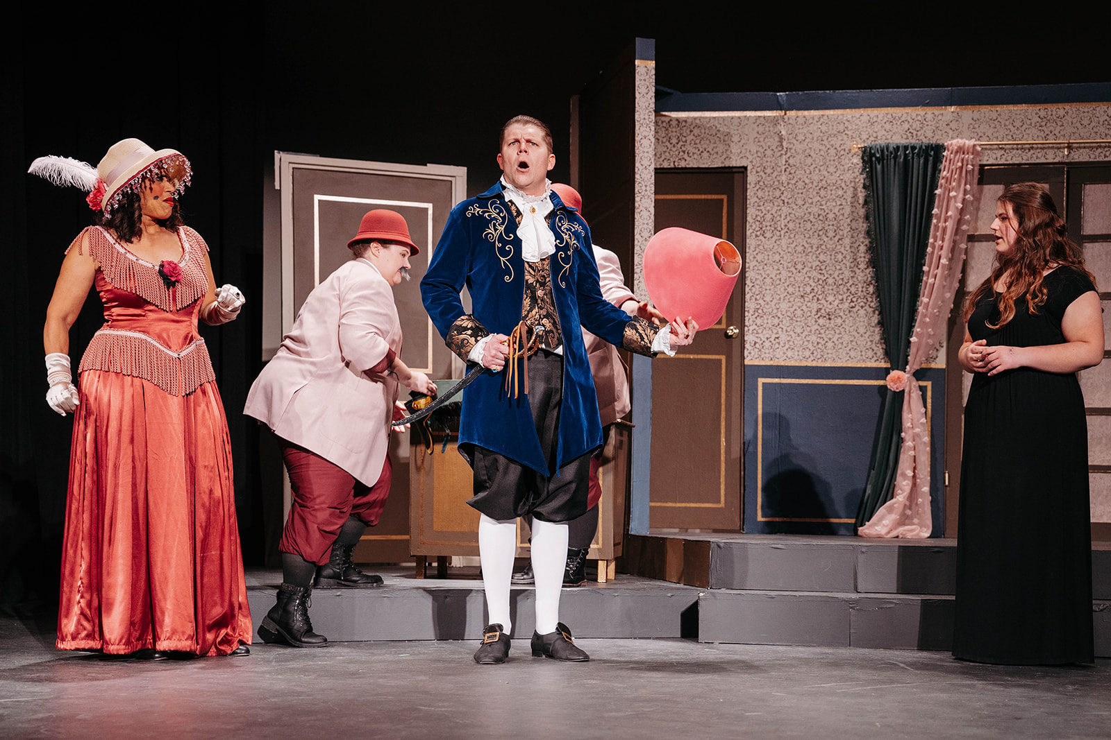 Rosetown Playhouse Three performers in period dress, including a woman in an ornate pink gown with feathered hat gesturing dramatically, a man in the blue coat looking surprised, and a woman in dark clothing observing.