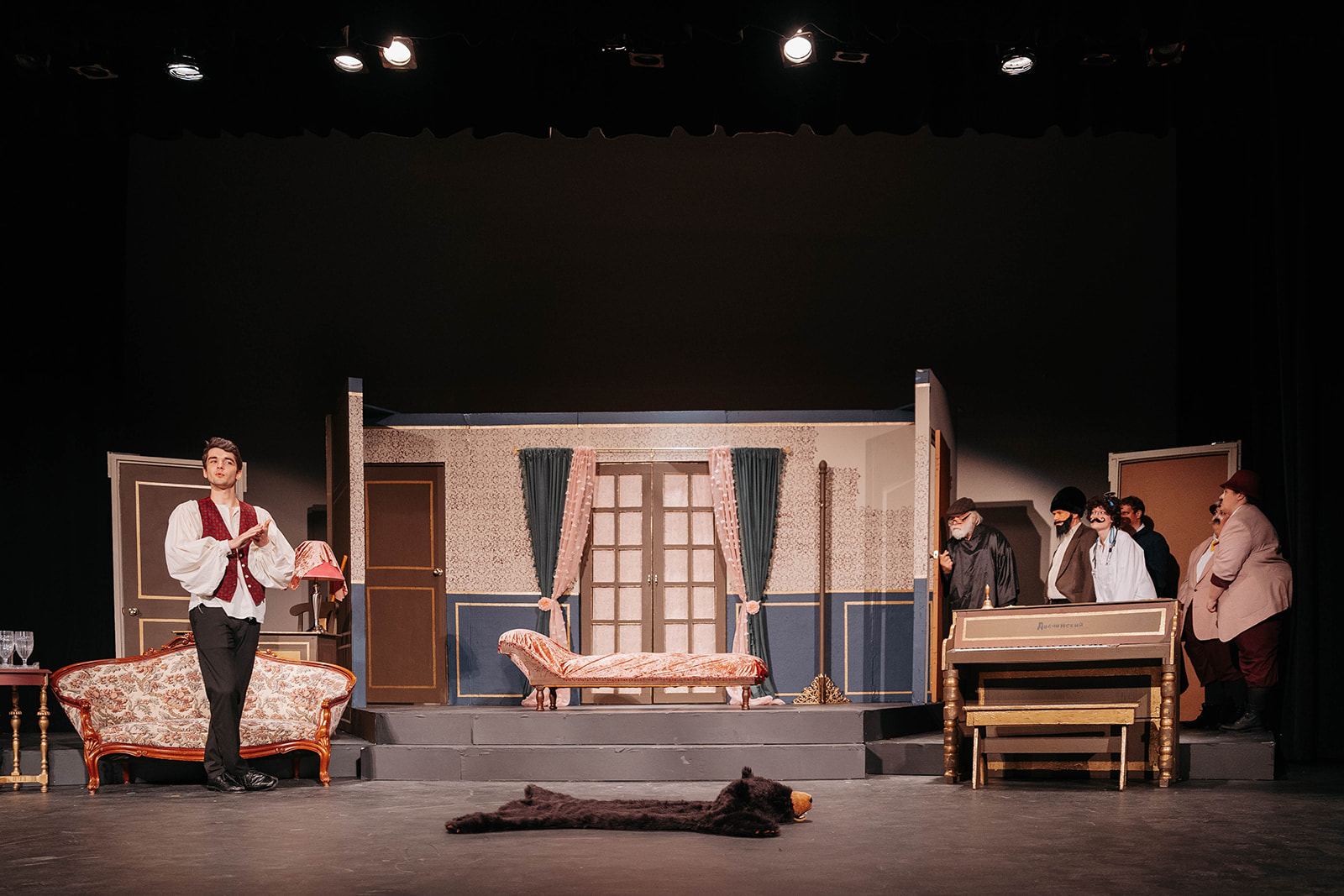 Rosetown Playhouse A lone performer in a vest stands downstage while other cast members in dark clothing lurk behind the set pieces in shadow.