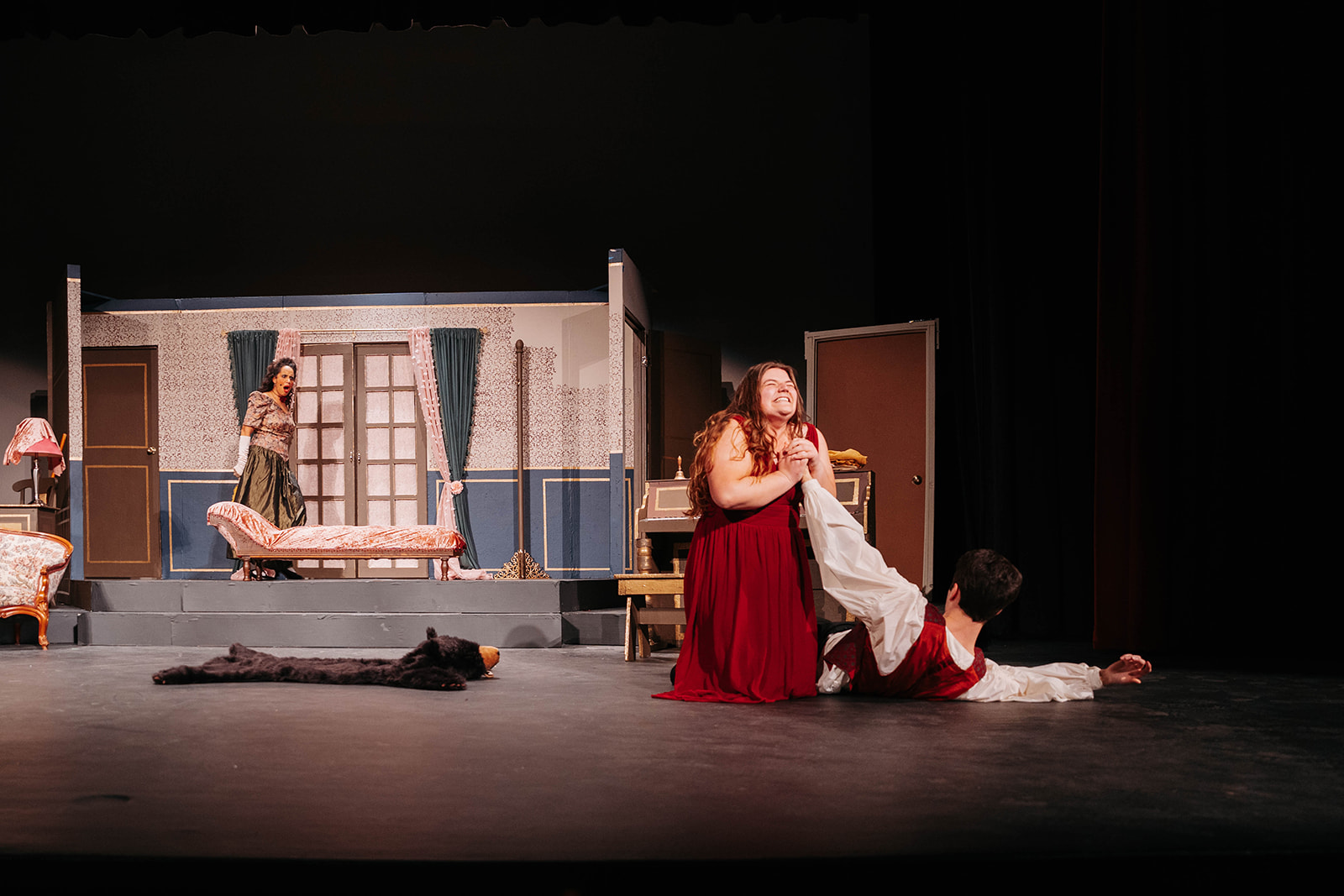 Rosetown Playhouse Two performers in an emotional scene, with one in a deep red gown clutching at another performer who kneels before them on the stage floor.