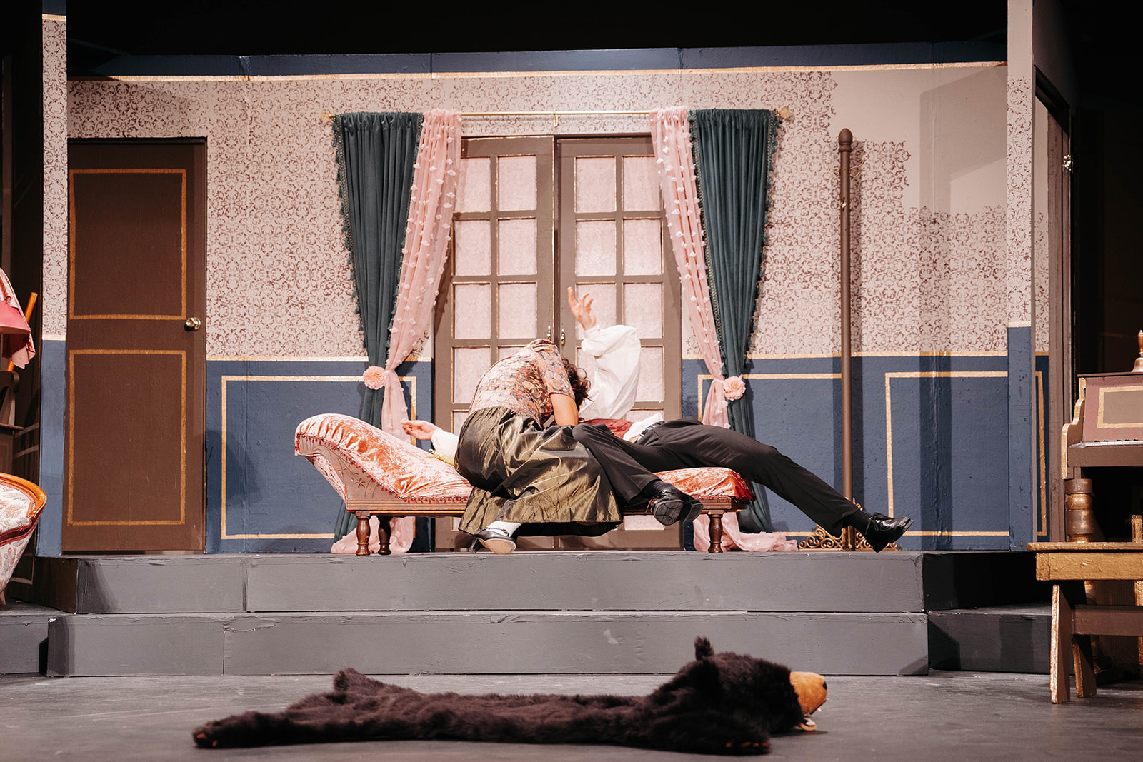 Rosetown Playhouse A comedic moment showing a performer lying dramatically across a chaise lounge in an exaggerated pose while being attended to by another in dark clothing.