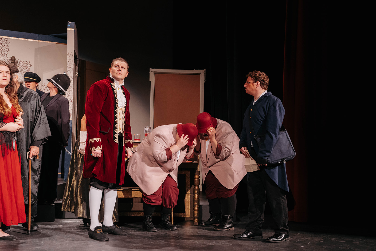 Rosetown Playhouse scene showing the cast in various positions, with officials bowing or covering their faces in shame while a character in a rich red velvet coat stands prominently and another in blue observes.