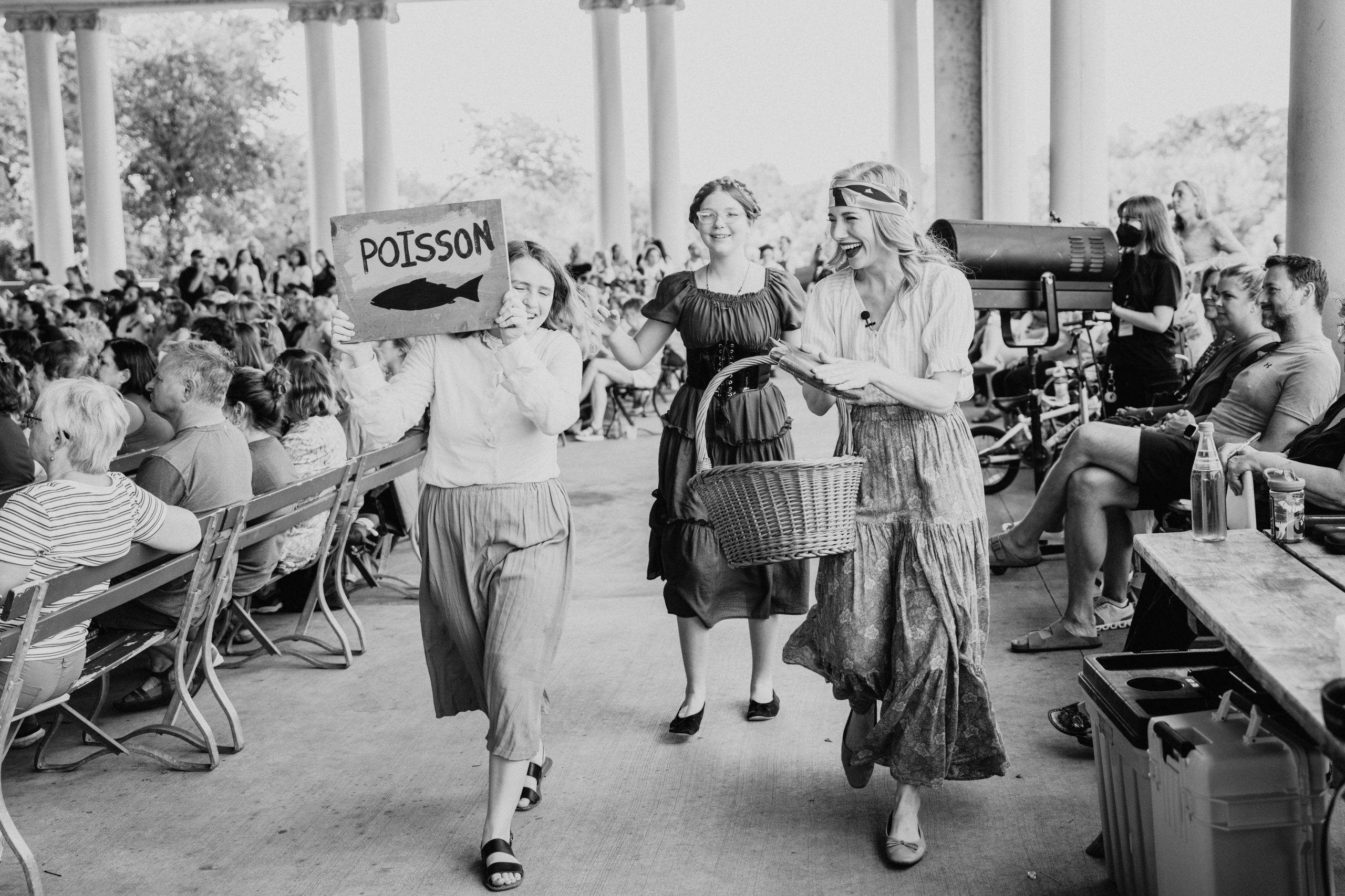 BeautyBeast_01 Black-and-white photo of three costumed performers walking up the aisle of an outdoor pavilion past a seated audience; one holds a hand-painted "Poisson" sign with a fish, another carries a wicker basket, all laughing mid-scene.