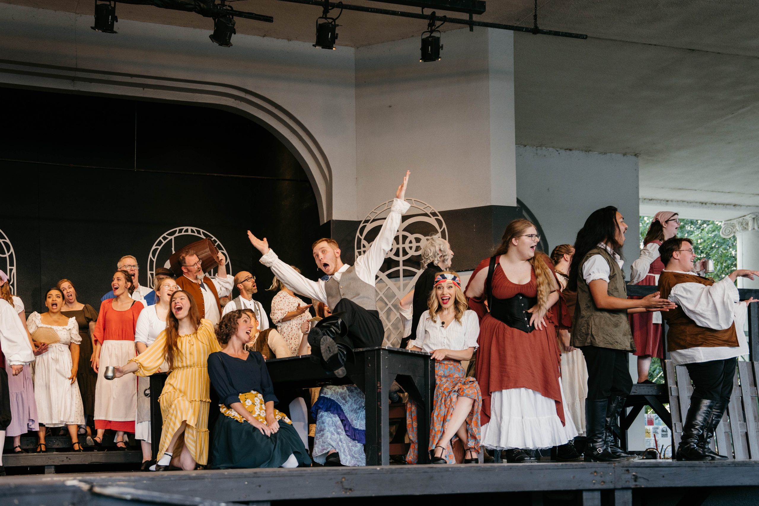 BeautyBeast_02 The full cast of Rosetown Playhouse's Beauty and the Beast performs an energetic group number on an outdoor pavilion stage, one actor leaping with his legs kicked up over a table as the ensemble sings with arms raised.
