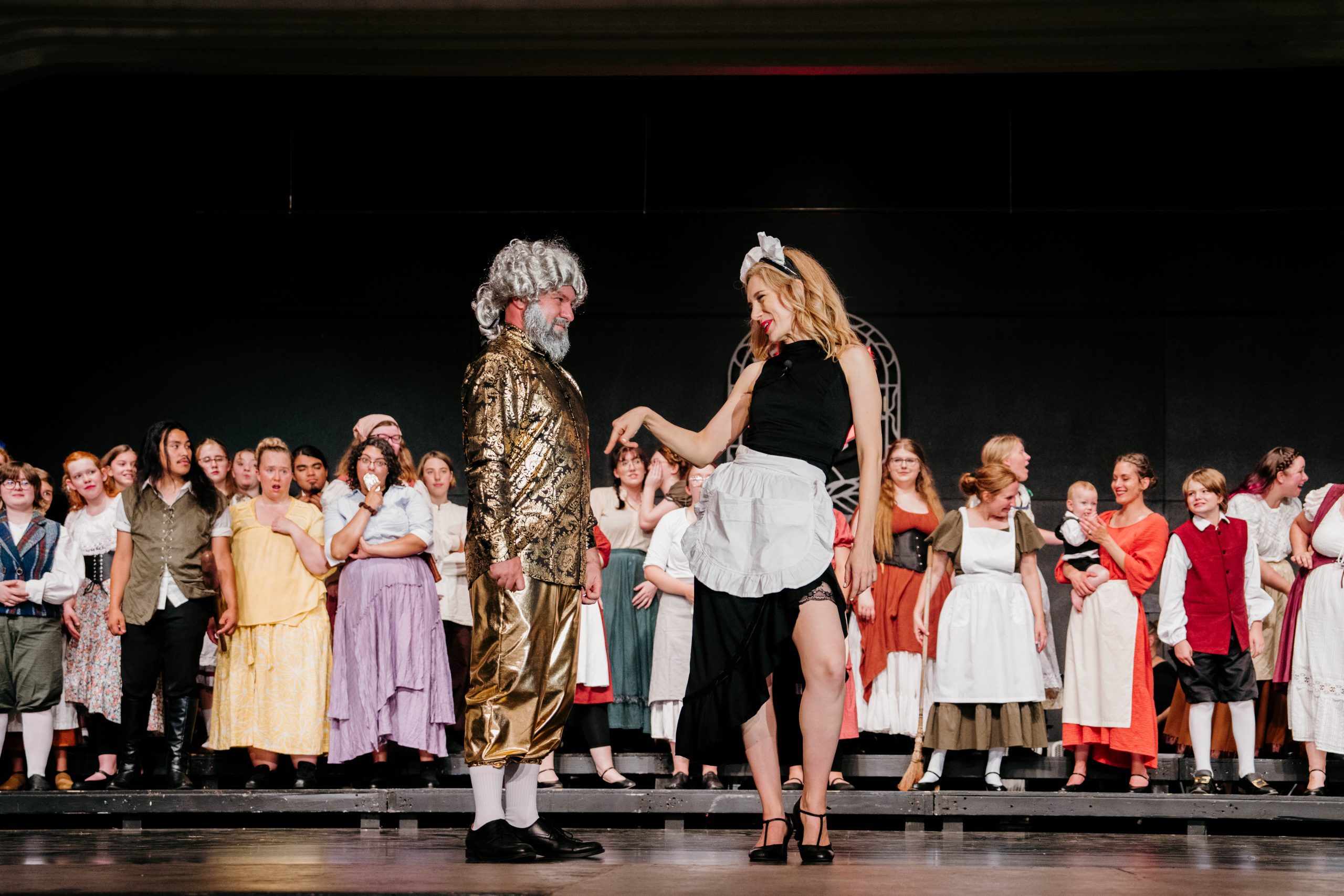 BeautyBeast_03 Two performers dance center stage in Rosetown Playhouse's Beauty and the Beast — a man in a gold brocade coat, breeches, and powdered wig facing a smiling woman in a black dress and white ruffled apron — while a large costumed ensemble in colorful period dress watches from behind.